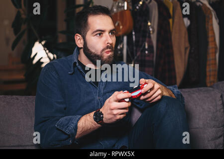 Porträt eines jungen Mannes spielen Videospiele, die sich auf die Fortschritte, die auf dem Rand seines Sitzes auf der Wohnzimmer Couch. Stockfoto