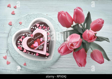 Valentine Herz Kuchen mit Schokolade, Zucker Dekorationen und Text 'Happy Valeitine" und ein Bündel von rosa Tulpen Stockfoto