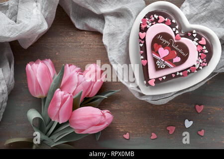 Valentine Herz Kuchen mit Text "Happy Valentine". Draufsicht auf dunklem Hintergrund mit rosa Tulpen und Bettwäsche Handtuch. Stockfoto