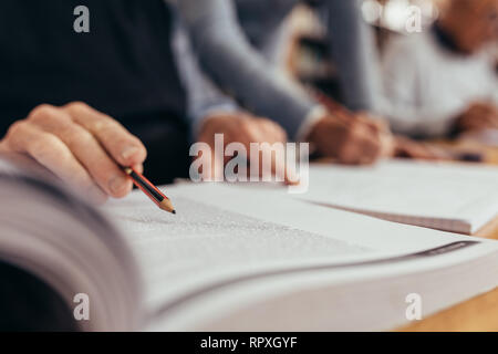 Nahaufnahme der Hand der Mann mit einem Bleistift und bei der Text in einem Buch zeigt. 7/8 Schuß einer Person ein Buch lesen, einen Bleistift in einem Cla sitzen Stockfoto