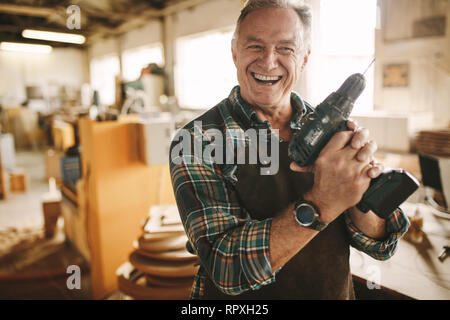 Lächelnd senior Tischler holding Bohrmaschine gegen Workshop. Reife männliche Arbeiter lächelte zuversichtlich in die Kamera halten Bohrmaschine an seinem Stockfoto