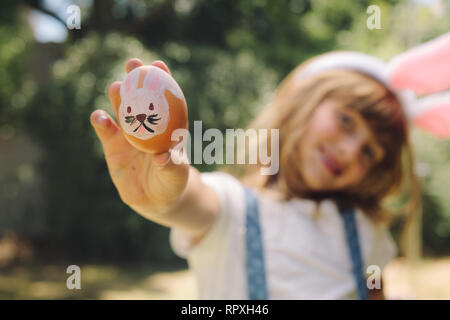 Lächelnde Mädchen Holding eine bemalte Ostereier in der Hand. Mädchen Anzeige ein Osterei mit Hase bemalt. Stockfoto