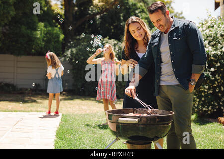 Glücklich, Mann und Frau, die Grillen gemeinsam in ihrem Hinterhof. Mädchen spielen mit Seifenblasen, während ihre Eltern Essen zu kochen für eine besondere Gelegenheit Stockfoto