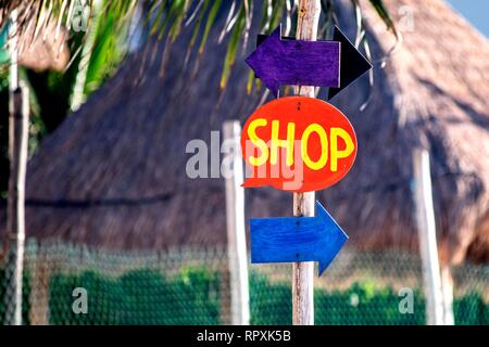Hand Made Sprechblase Form Shop anmelden und Richtungspfeile auf einen Beitrag in einer tropischen Anlage mit strohgedeckten Hütten im Hintergrund. Stockfoto