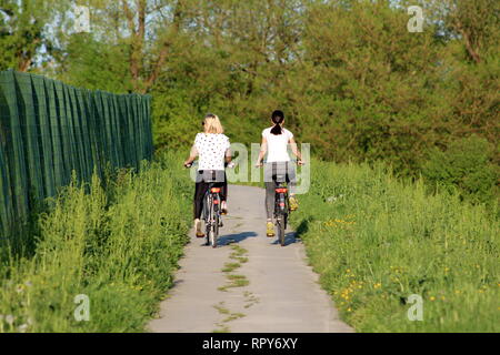 Zwei schöne junge Mädchen am Sport Outfit, ihre Fahrräder auf asphaltierten Feldweg, der mit hohen, grünen Gras und Wald Vegetation umgeben Stockfoto