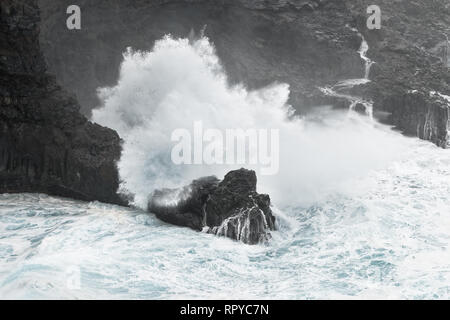 Eine Welle bricht bei stürmischem Wetter auf einer felsigen Küste mit einem kleinen Bucht, Gischt spritzt weit oben, aus dem Felsen, Wasser läuft wieder zurück ins Meer - Ort: Spa Stockfoto