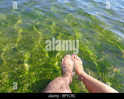 Nahaufnahme eines männlichen Bein und eine weibliche Bein vom Knie bis zu den Füßen in glasklaren grünen seichten Meer Wasser. Zweisamkeit. Stockfoto