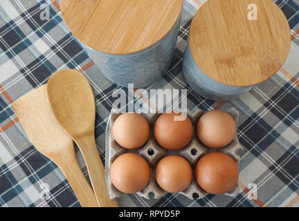 Sechs rohe Eier im Karton auf dem Küchentisch top Aussicht, mit Löffel aus Holz und Küche Behälter für Lebensmittel Ansicht von oben. Kochen und Backen Konzept Stockfoto