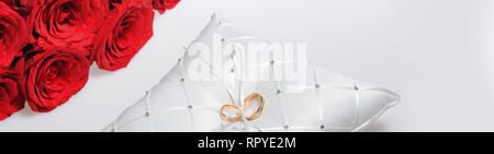 Schließen Sie zwei Hochzeit goldene Ringe liegend auf Weiße weiche Kissen und ein Strauß roter Rosen über weiß Panoramablick Hintergrund, Copyspace für Text. Die Ehe. Stockfoto