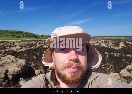 Selfie eines Jungen bärtigen Mann in einer Sun Hut auf einem Sommer Tag der Erkundung der Küste entlang der zerklüfteten Küste von Fife. Schottland, Großbritannien. Stockfoto