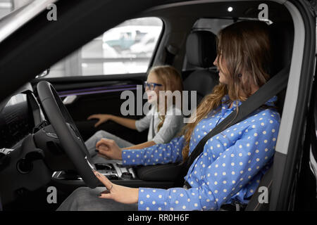 Frau sitzt im Fahrersitz mit Sicherheitsgurt, halten sich an den Händen am Lenkrad. Ihre Tochter vor Sitz sitzt, lächelnd, entfernt. Familie testen und beobachten neues Fahrzeug. Stockfoto