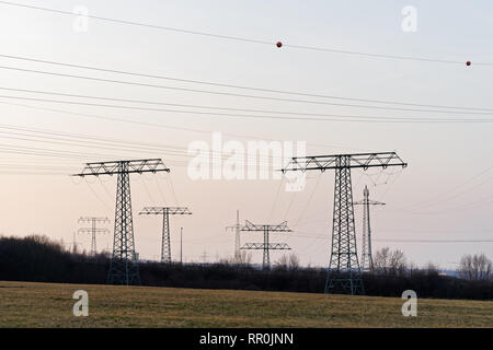 Große Sammlung von Strommasten in verschiedenen Arten mit Kabeln, stehen sie gestaffelt als Silhouetten vor einem leicht gefärbten Abendhimmel Stockfoto