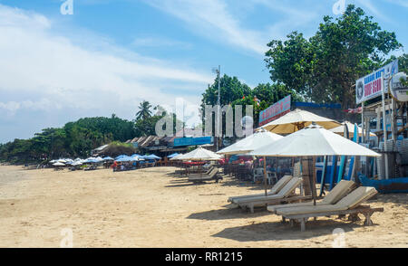 Liegestühle und Sonnenschirme am Strand vor eine Reihe von Fischrestaurants Jimbaran Bay Bali Indonesien. Stockfoto