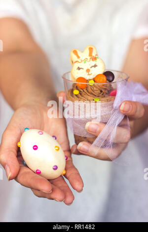 Woman's Hände halten transparente Schale aus Kunststoff mit Mousse au Chocolat in der einen Hand und gestaltet Ostereier in den anderen Stockfoto