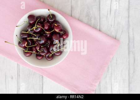 Kirschen in ein weißes Porzellan Schüssel auf rosa Stoff auf einem weißen Laufbelag Hintergrund. Ansicht von oben. Stockfoto