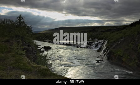 Längere Exposition von Wasserfall Hraunfossar am Fluss Hvita mit bedecktem Himmel mit einigen Sonnenlicht durch die Wolken. Island. Stockfoto