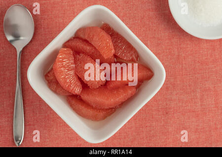 Rote Grapefruit in einem quadratischen weißen Schale mit einem Löffel und eine kleine runde Schüssel mit Zucker auf einer Koralle Hintergrund Stockfoto