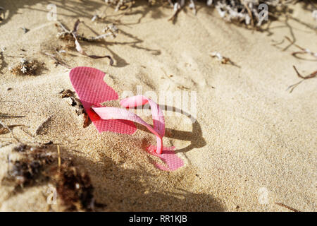 Eine einzelne rosa Flipflop Badeschuhe liegt auf einem sandigen Strand menschenleer, es ist teilweise mit Sand bedeckt, zusammen mit Algen und andere Pflanze bleibt, st Stockfoto