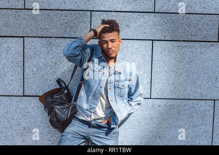 Freestyle. Mulatte Kerl mit Rucksack stehend an der Wand zu berühren, das Haar Kamera verspielt isoliert Stockfoto