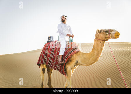 Stattlich im Nahen und Mittleren Osten Mann mit kandura und gatra reiten auf einem Kamel in der Wüste Stockfoto