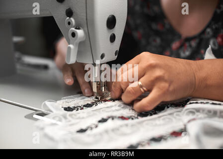 In der Nähe der weiblichen Näherin Hände verbinden weißen Stoff details mit Blumen bunte Stickerei mit langen Streifen der Spitze auf moderne Nähmaschine. Modische Kleidung Fertigungskonzept. Stockfoto