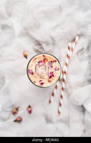 Eiskaffee mit Rose und Kardamom in ein hohes Glas mit getrockneten Rosen und Strohhalme auf weißer und grauer Seide Hintergrund umgeben. Ansicht von oben mit der Kopie Raum fo Stockfoto