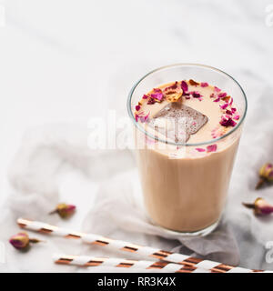Eiskaffee mit Rose und Kardamom in ein hohes Glas mit getrockneten Rosen und Strohhalme auf weißer und grauer Seide Hintergrund umgeben. Kopieren Sie Platz für Text. Platz Stockfoto