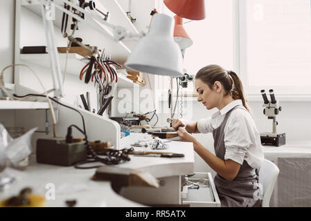 Besetzt. Seitenansicht des jungen weiblichen Juwelier arbeiten an einer neuen Schmuck Produkt an Ihrer Werkbank. Schmuck. Business. Schmuck Werkstatt. Stockfoto