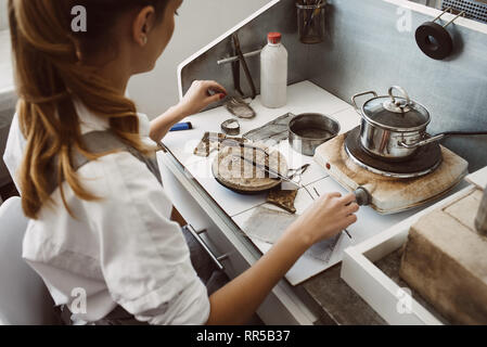 Nachdenken über neue Produkt. Seitenansicht des jungen weiblichen Juwelier bei ihrem Schmuck, Werkstatt und denken, welche Werkzeuge Sie für eine neue Verwendung sitzen Stockfoto