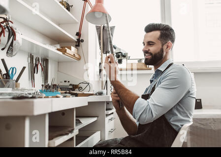 Bei der Arbeit zufrieden. Porträt der jungen und freundlichen männlichen Juwelier Kontrolle ring Größe mit Spezialwerkzeug in der Werkstatt. Schmuck Werkstatt. Juwelier Tools Stockfoto