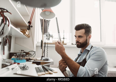 Es ist keine leichte Arbeit. Portrait von jungen männlichen Juwelier Kontrolle ring Größe mit Spezialwerkzeug in der Werkstatt. Schmuck Werkstatt. Juwelier Tools Stockfoto