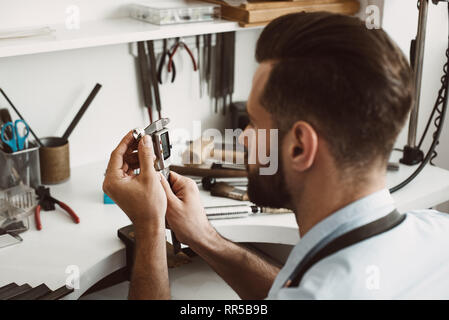 Genaue Größe. Nahaufnahme Foto eines jungen männlichen Juwelier messen Ring mit einem Werkzeug in der Werkstatt. Schmuck Herstellung Konzept. Schmuck Werkstatt. Mas Stockfoto