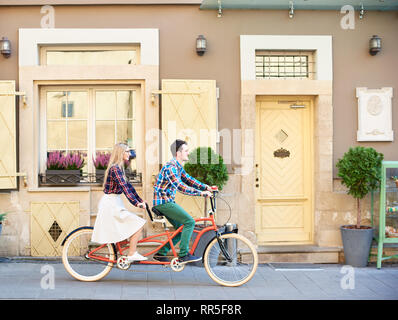 Seitenansicht der aktiven touristische Paar in Freizeitkleidung, bärtigen Mann und die blonde Frau in den Brillen Fahrrad tandem Fahrrad entlang Leer gepflasterten Gehweg auf die hellen sonnigen Sommertag nach Stadt Wohnhäuser. Stockfoto