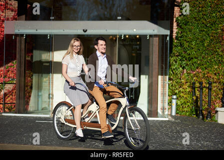 Junge lächelnde Paar, gut aussehenden bärtigen Mann und attraktive Langhaarige blonde Frau radfahren Tandem Bike auf sonnigen Tag der offenen Glastür von Backstein Gebäude voll mit roten Efeu Blätter überwuchert. Stockfoto