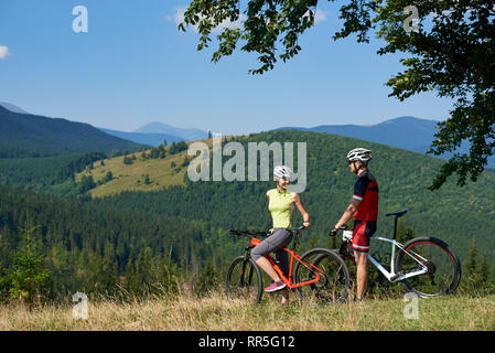 Happy biker Paar, Mann und Frau, mit Fahrrädern auf grasbewachsenen Hügel unter der großen grünen Zweig der Baumstruktur, lächelnd und Miteinander auf der Suche nach sonnigen Sommertag. Herrliche Aussicht auf die Bergkette im Hintergrund Stockfoto
