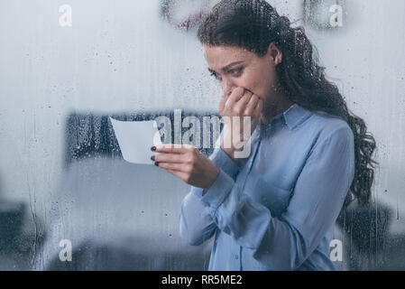 Trauernde Frau mit Foto, die den Mund mit der Hand und zu Hause weinen durch Fenster mit Regentropfen Stockfoto