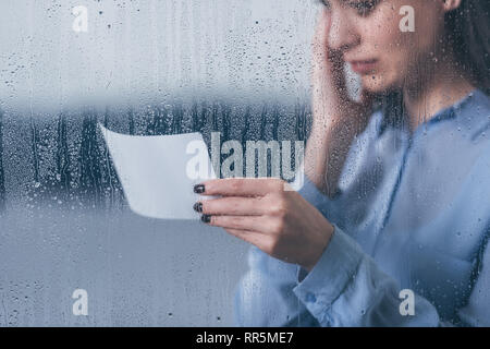 7/8-Ansicht der Trauernde Frau mit Foto und weinend durch Fenster mit Regentropfen Stockfoto