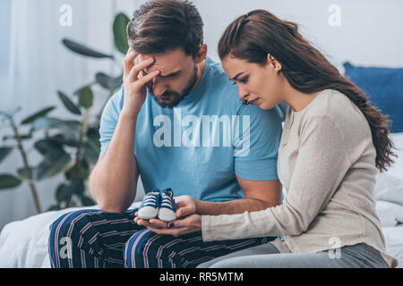 Selektiver Fokus der trauernde Eltern sitzen auf dem Bett und Holding Baby Schuhe zu Hause Stockfoto