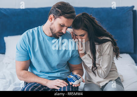 Selektiver Fokus der trauernde Eltern sitzen auf dem Bett und Holding Baby Schuhe zu Hause Stockfoto