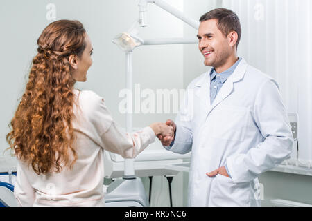 Fröhliche Zahnarzt stehend mit Hand in der Tasche und Händeschütteln mit weiblichen Patienten Stockfoto