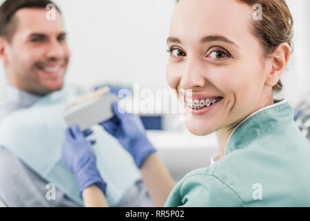 Selektiver Fokus fröhliche Zahnarzt mit Klammern an den Zähnen lächelnd, während sie Farbpalette in der Nähe von Patienten Stockfoto