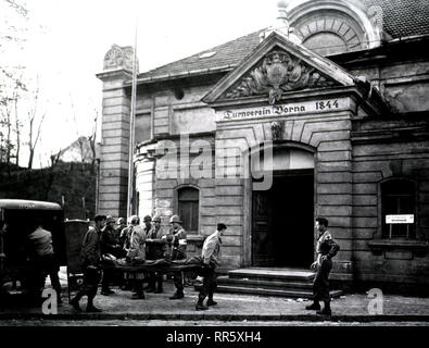 Ein Fahrzeug mit der hinteren Türen geöffnet sitzt vor einem Gebäude aus dem 19. Jahrhundert, in ein Lazarett umgewandelt wurde. (Unter dem Giebel ist eingraviert: "Turnverein Borna 1844.') Zwei Soldaten tragen eine verletzte Soldaten auf einer Bahre aus dem Fahrzeug auf die offene Tür eines Gebäudes. Stockfoto