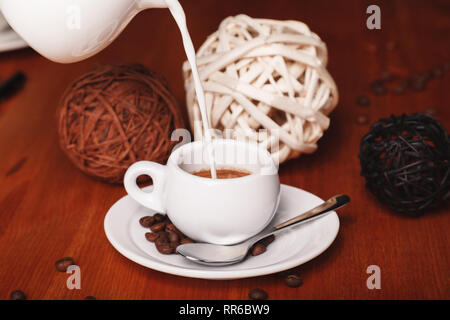 Gießen Milch aus der Milchmann in eine kleine Tasse Espresso auf einem hölzernen Hintergrund. Das Konzept der Kaffeepausen und Kaffee Stockfoto