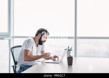 Portrait eines bärtigen dunkelhaarigen Mann sitzen, der mit einem weißen Büro und auf seinem Laptop Bildschirm, während Sie telefonieren. Seine Tasse Kaffee wird in f Stockfoto