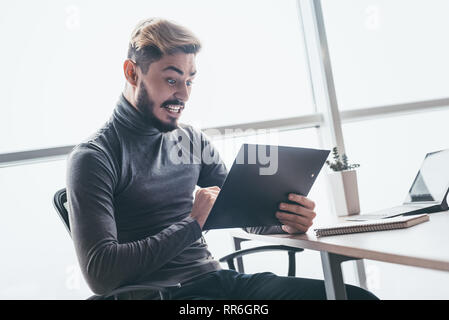 Jungen betonte Unternehmer sitzen mit Diagrammen in seiner Hand mit entsetzten Gesichtsausdruck. Überrascht Geschäftsmann auf Ergebnisse, besorgt und amaz Stockfoto