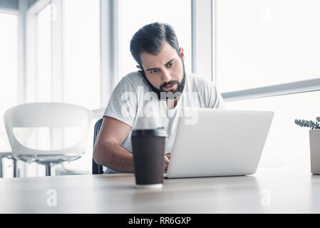 Portrait eines bärtigen dunkelhaarigen Mann sitzen, der mit einem weißen Büro und auf seinem Laptop Bildschirm, während Sie telefonieren. Seine Tasse Kaffee wird in f Stockfoto