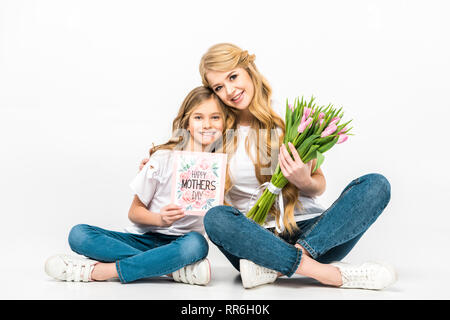 Mutter mit Blumenstrauß aus rosa Tulpen und Tochter mit glücklichen Muttertag Grußkarte auf dem Boden Sitzen mit gekreuzten Beinen auf weißem Hintergrund Stockfoto