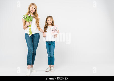Lächelnde Mutter mit Blumenstrauß aus rosa Tulpen und lächelnden Tochter mit glücklichen Muttertag Grußkarte halten sich an den Händen und Kamera schaut auf Weiß b Stockfoto
