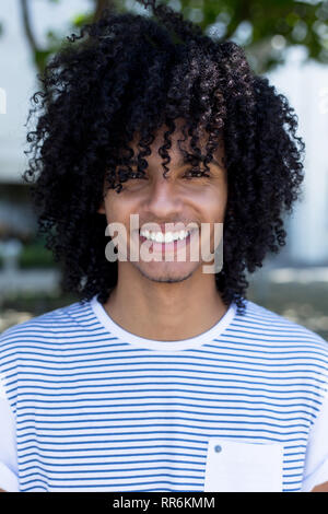 Portrait von Lachen lateinamerikanischen junger erwachsener Mann mit Langen lockigen Haar im Freien in der Stadt Stockfoto