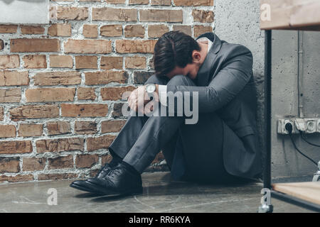 Trauriger Mann im grauen Anzug sitzen auf dem Boden und umarmen Knien auf Braun strukturierten Hintergrund in Zimmer, trauernde Unordnung Konzept Stockfoto